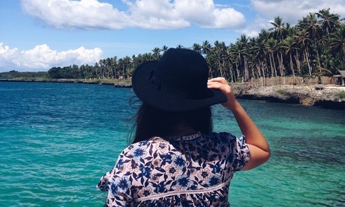 брюнетка в шляпе со спины смотрит вдаль на море на берегу много пальм