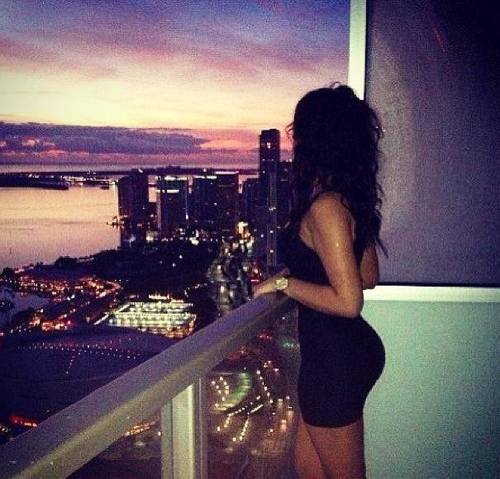 брюнетка в черном платье на балконе смотрит на ночной мегаполис