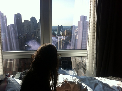брюнетка со спины без лица у окна смотрит на город с высотки в постели