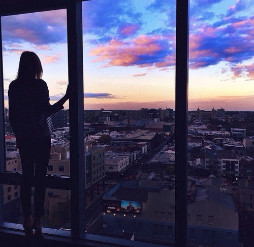 девушка у панорамного окна на заходе солнца вечерний город без лица со спины