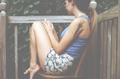 дівчина з татуюванням пера на плечі сидить на стільці на ганку