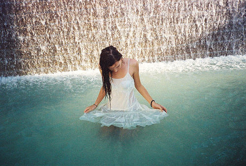 дівчина з чорним волоссям в легкому білому платті купається у водоспаді