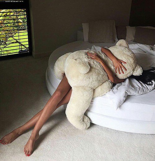 брюнетка лежит на кровати с огромным плюшевым медведем