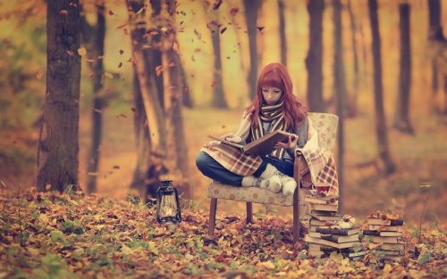 девушка укуталась в плед с книгами на лавочке среди осеннего леса с фонариком