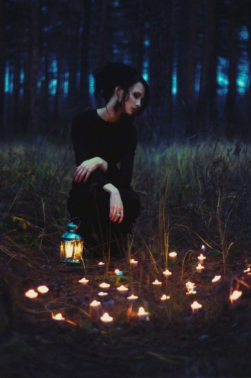 брюнетка в черном платье в ночном лесу среди свечей и фонаря