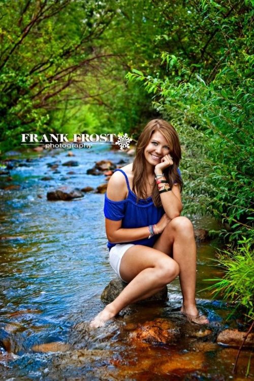 девушка в синей футболке и белых шортах сидит на камне в реке летом среди зеленых деревьев