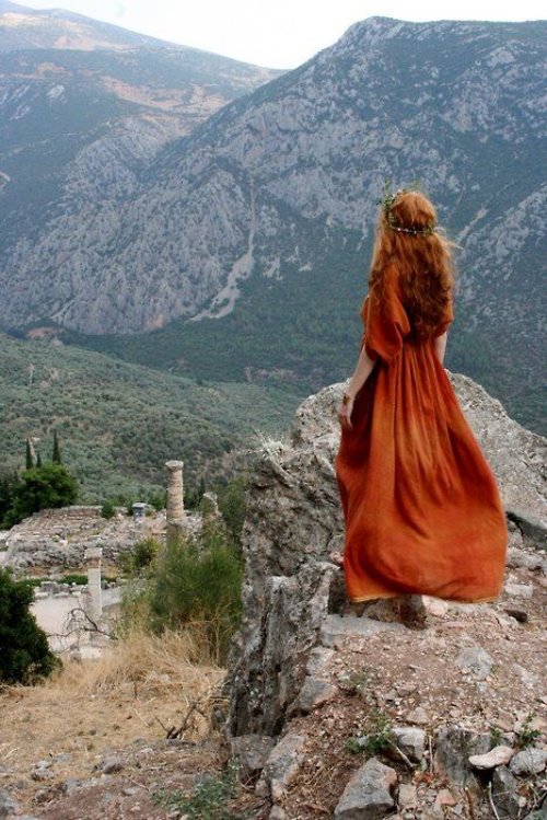 рыжая девушка в длинном красном платье на вершине горы смотрит вдаль со спины