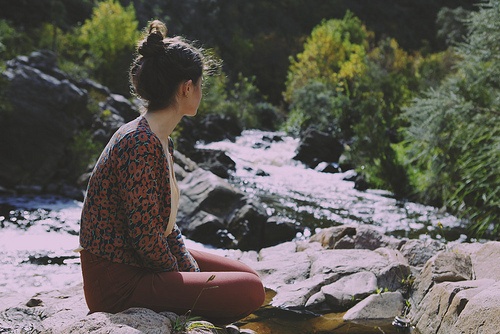 девушка с пучком в брюках и блузе в цветочек сидит на камне у реки
