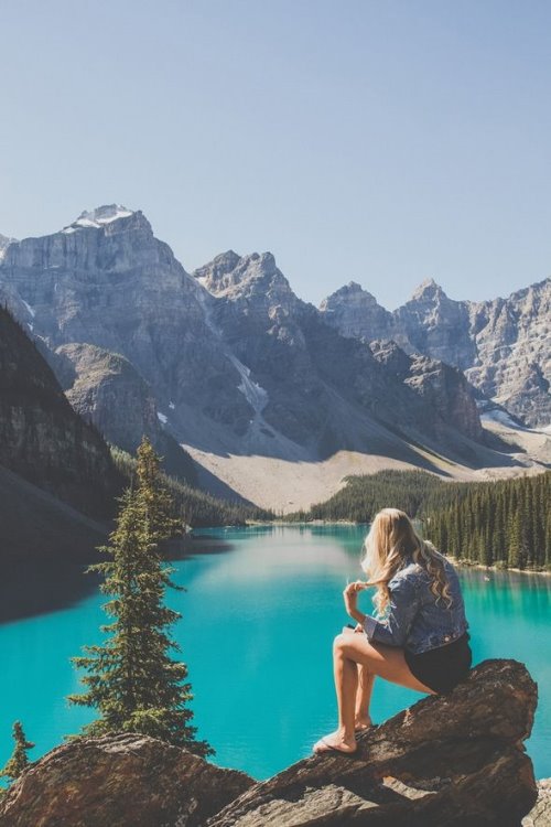 блондинка с локонами со спины сидит на камне смотря на голубое озеро в горах