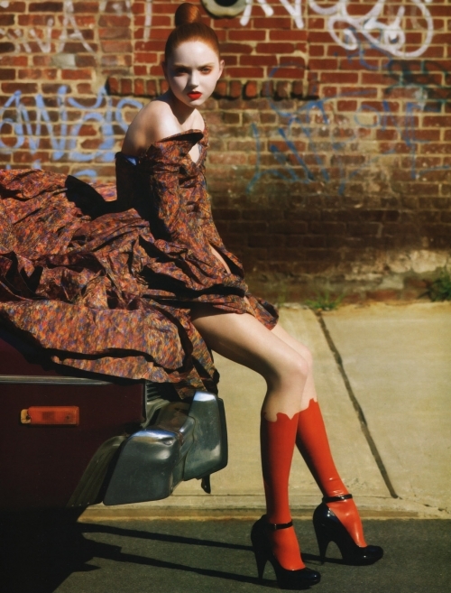 рыжая девушка с пучком на капоте автомобиля на фоне кирпичной стены
