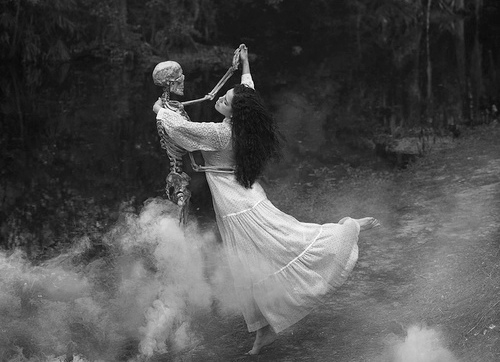 брюнетка в длинном белом платье танцует со скелетом ночью в лесу