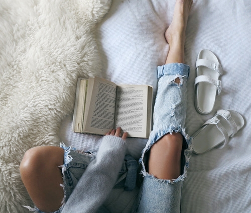 девушка читает книгу в кровати со шкуркой мериноса в свитере и порванных на коленях джинсах