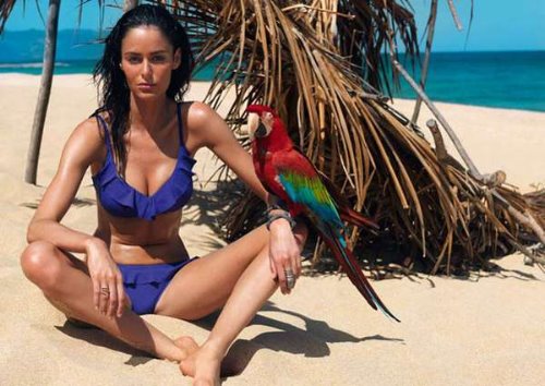брюнетка в синем купальнике с рюшами с попугаем на руке сидит на пляже под пальмой