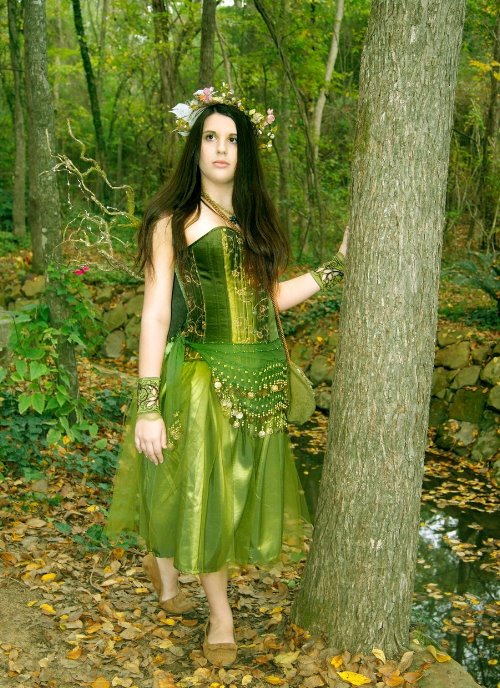 полная девушка в образе лесной нимфы фотографируется под деревом в лесу в шелковом зеленом платье