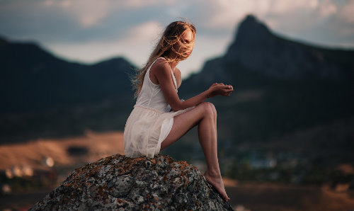 светловолосая девушка в белом сарафане сидит на камне в горах
