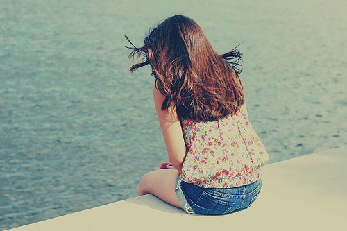 девушка спиной сидит в джинсовых шортах и цветочной блузе на пирсе любуясь морем ветер треплет волосы