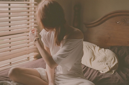 девушка с хвостиком смотрит в окно сквозь жалюзи сидя на кровати