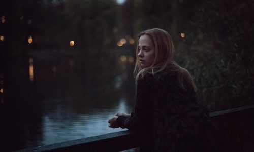блондинка облокотилась на мост и смотрит на воду со спины вечером