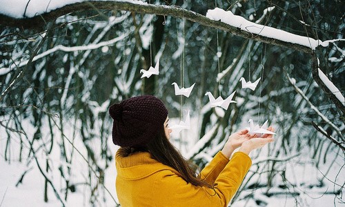 девушка в желтой куртке смотрит на оригами в зимнем снежном лесу