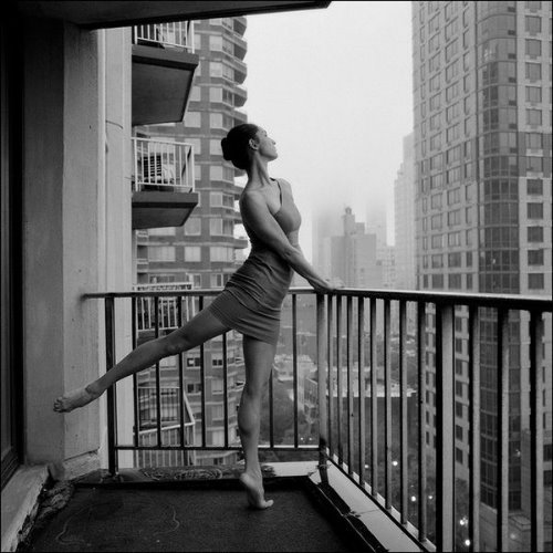 девушка машет ногой в платье с оголенным плечом на балконе