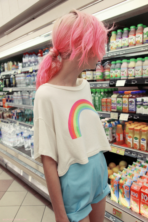 сутулая девушка в белой футболке с радугой и коротких голубых шортах бродит по магазину выбирая сок