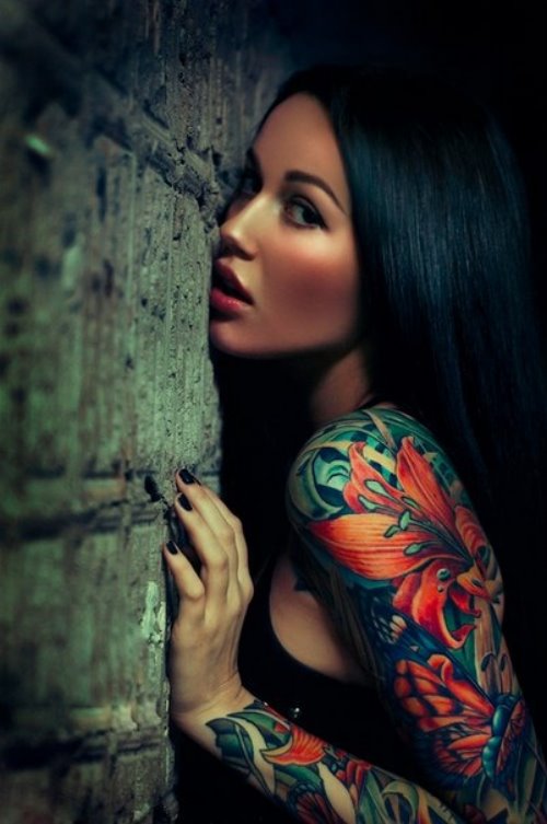 Брюнетка с черными ногтями и яркой татуировкой оранжевая лилия на руке прижалась щекой к каменной стене.