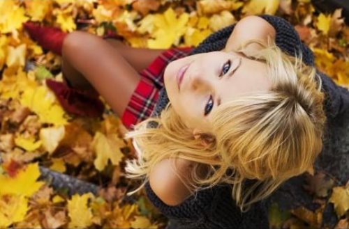 блондинка в короткой клетчатой юбке в красных туфельках сидит на желтых осенних листьях и смотрит вверх на фотографа