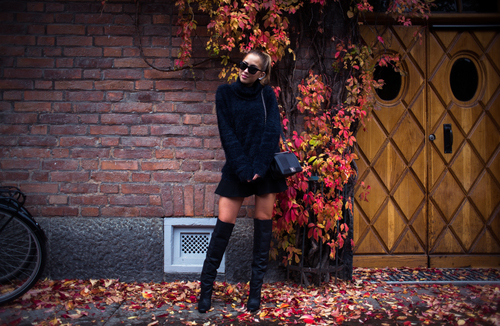 девушка в черном в высоких сапогах с хвостом и сумкой позирует на фоне стены и дикого красного винограда осенью