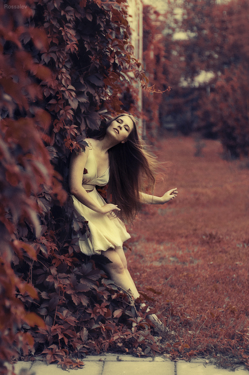 Девушка с длинными волосами с закрытыми глазами в белом платье стоит среди листьев дикого винограда.