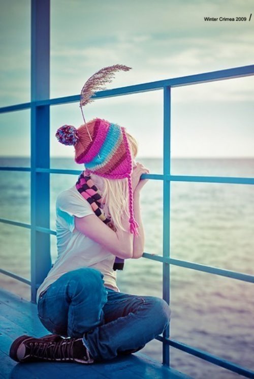 Девушка в яркой шапке с помпоном и полосатом шарфе сидит в кедах на пирсе и смотрит на холодное осеннее море.