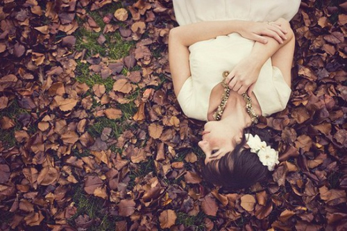 Девушка в белом платье с цветами в волосах держит свое ожерелье и смотрит на осенние коричневые листья в которых лежит.