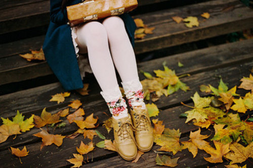 Ножки в белых колготках и цветочных гетрах в синем пальто и легкой белой юбочке сидят на деревянных ступеньках среди желтых осенних листьев.