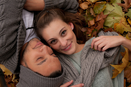 Влюбленная пара в серых свитерах лежат обнявшись в осенних листьях.