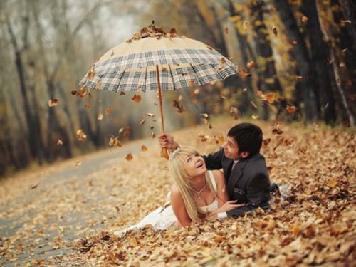 Влюбленная пара лежит на осенних листьях. Парень прячет девушку под клетчатым зонтом от листопада.