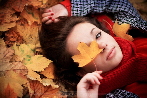 Шатенка в красном свитере, шарфе и с красным маникюром лежит в листьях и прикрывает правый глаз желтым кленовым листом.