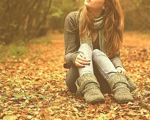 Девушка в джинсах и сером джемпере с шарфом сидит на осенних листьях и смотрит вверх.