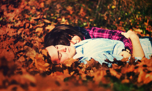 Влюбленная пара лежит в рыжих осенних листьях. Девушка в клетчатой рубашке, парень в голубой рубашке в белую полоску. Пара с закрытыми глазами.