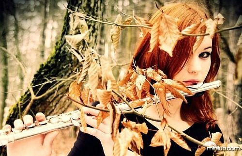 Рыжая девушка играет на флейте среди засохших осенних листьев в лесу.