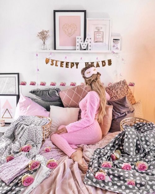 Девушка в розовой вязаной пижамке спиной с маской для сна сидит на кровати усыпанной розами