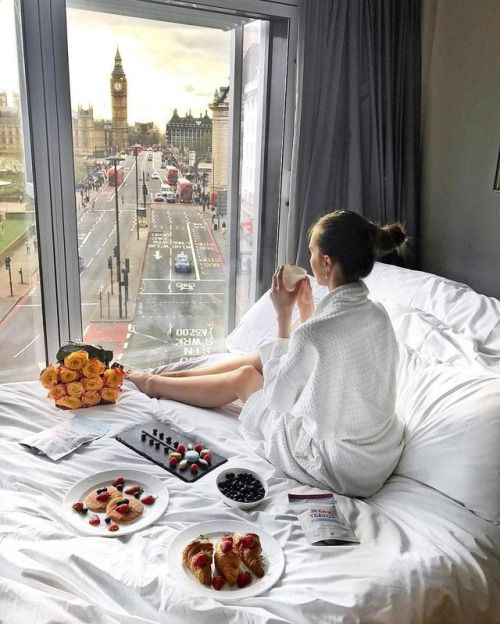 Девушка спиной в кровати с оладушками и круассанами смотрит в окно на утренний город