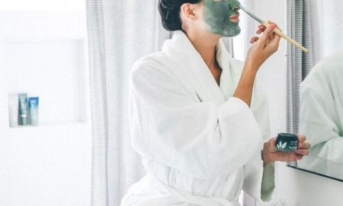 Девушка в ванной в халате наносит маску кисточкой перед зеркалом