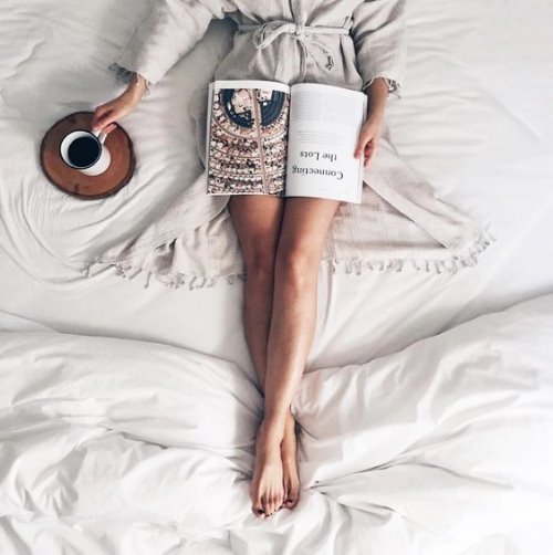 Девушка со стройными ногами лежит на кровати с книгой и чашкой свежезаваренного кофе