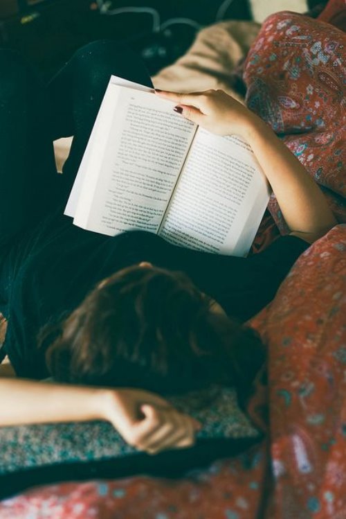 Девушка лежа на спине читает книгу облокотившись о диван
