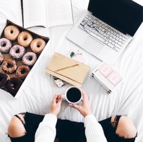 Девушка с пончиками и кружкой чая работает дома на ноутбуке сидя на кровати в рваных черных джинсах