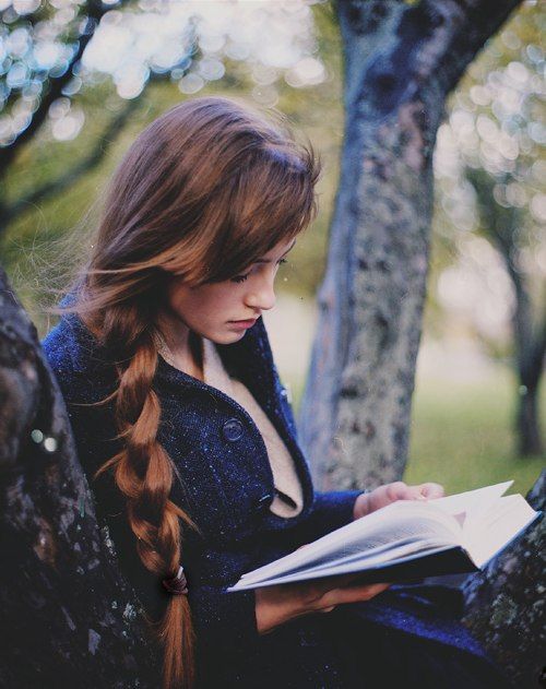 Девушка с косой в пальто читает книгу под деревом