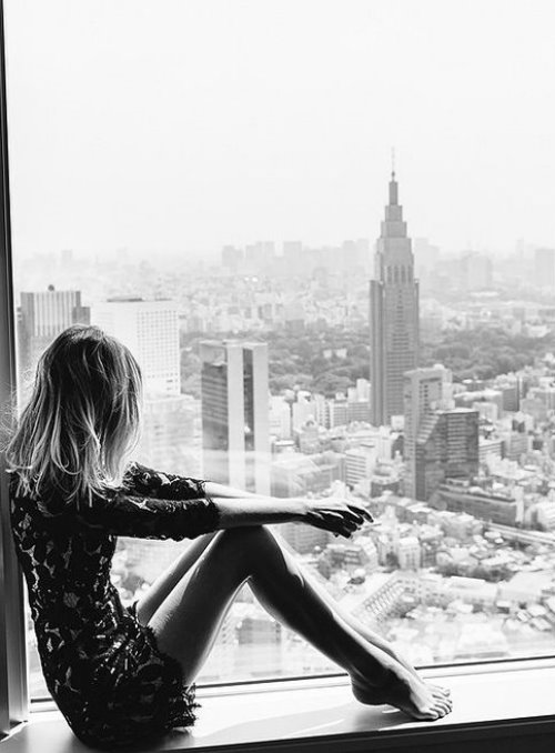 Блондинка смотрит свысока на город сидя на подоконнике