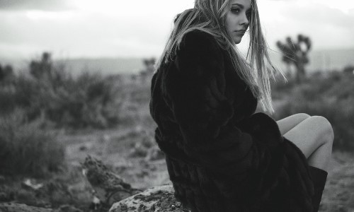 красивая блондинка в шубке и сапогах сидит на камне черное белое фото со спины