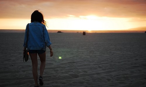 девушка в джинсовой короткой куртке и шортах со спины идёт по песку на закате