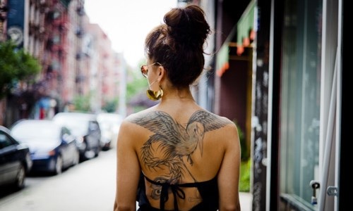 девушка с коричневыми волосами идет по городу среди машин с татуировкой на спине
